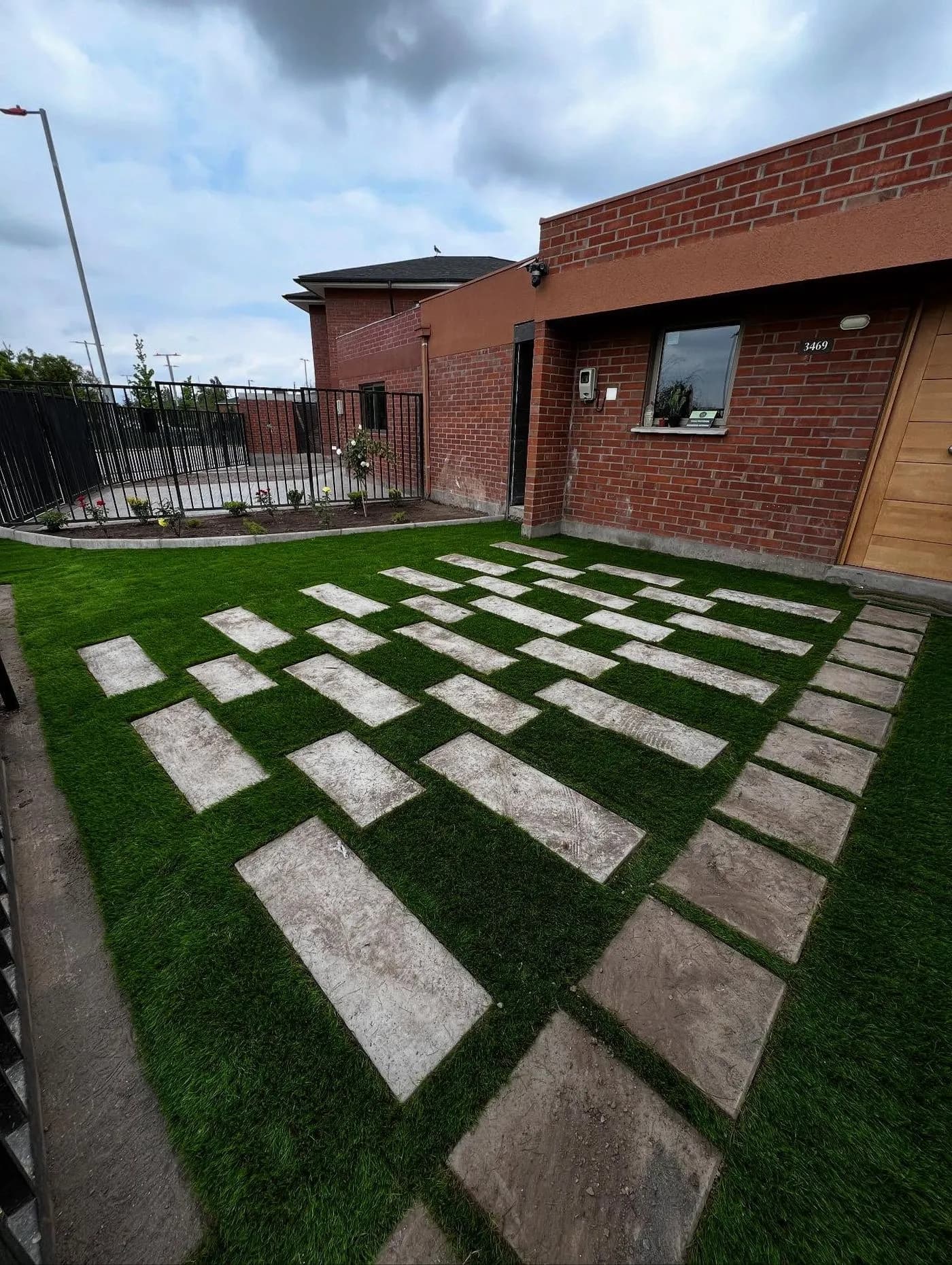 Jardín diseñado con baldosas rectangulares dispuestas en damero sobre el pasto, frente a una casa de ladrillo